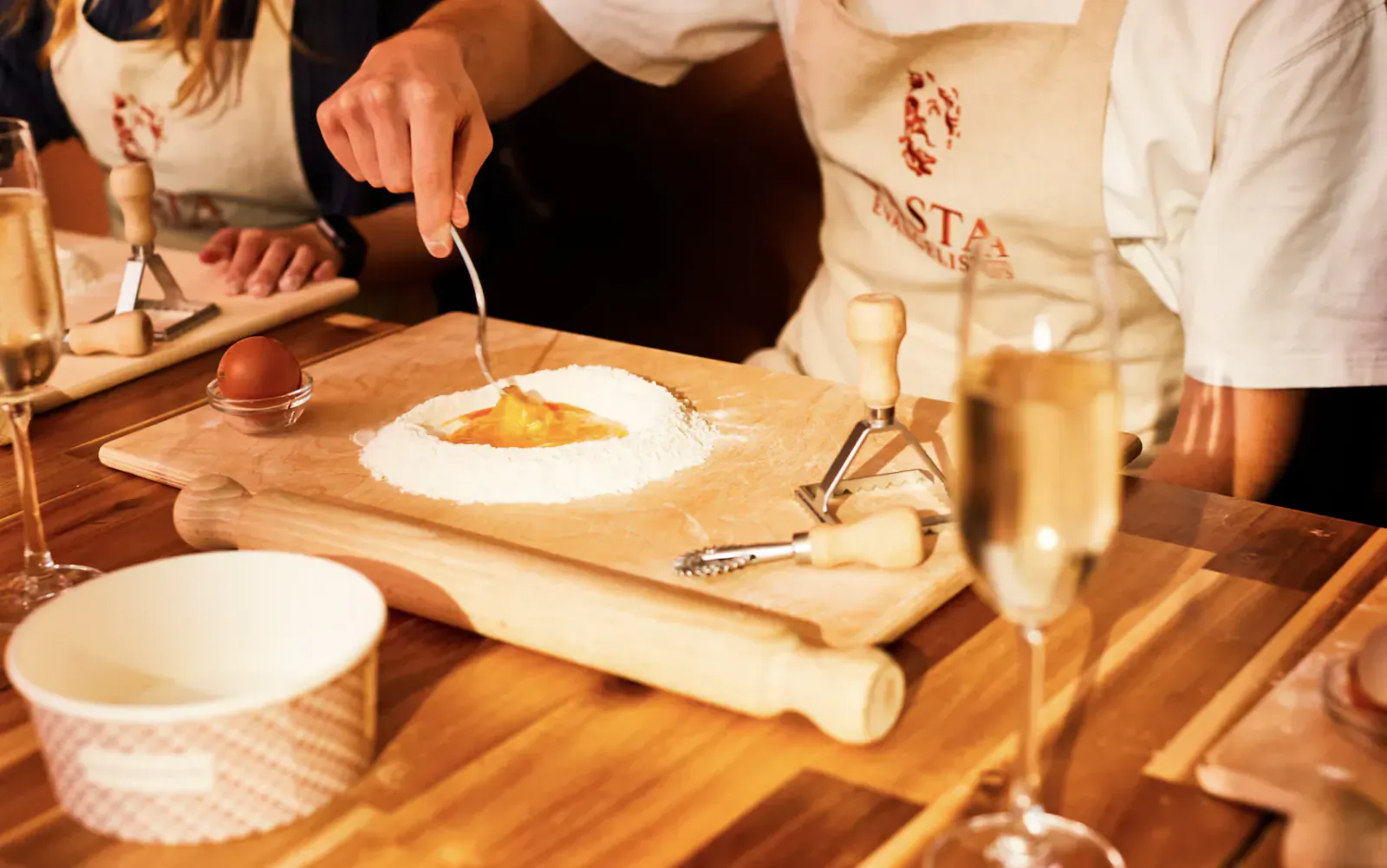 Pasta Evangelists Academy chefs in aprons making pasta dough with egg yolk on wooden board, rolling pin and champagne glasses nearby.