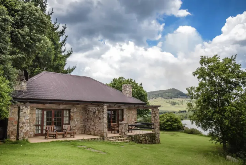 Stone cottage with veranda and chairs on grassy lawn by lake, surrounded by trees, hills under partly cloudy sky