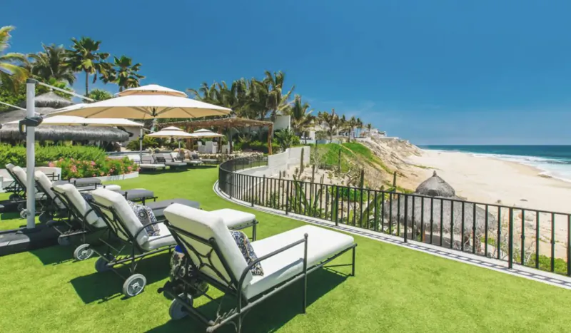 Luxury resort poolside lounge chairs on green lawn overlooking ocean beach, palm trees and umbrellas