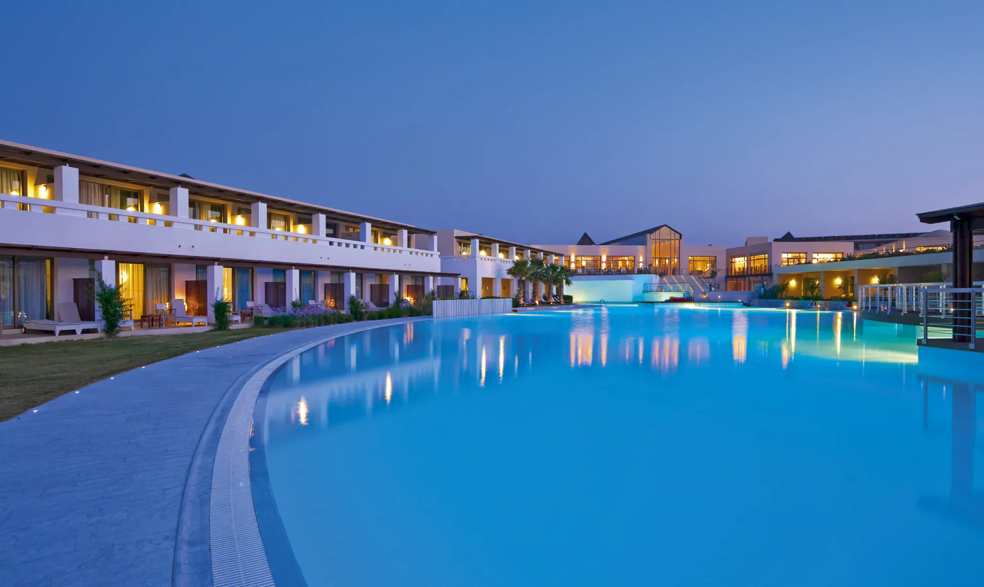 Twilight view of Giannoulis Cavo Spada Crete luxury resort with curved blue infinity pool, white buildings, and palm trees.