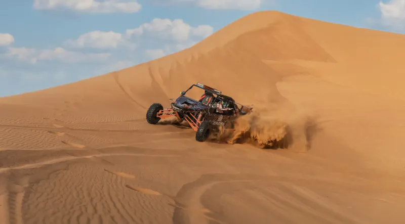 Orange Can-Am Maverick X3 dune buggy with riders speeding up sandy dune, kicking up dust in desert landscape