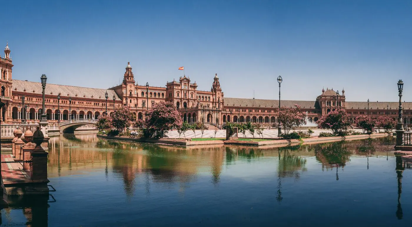 Panoramic view of Seville's Plaza de España with red brick architecture, arches, towers, and reflections in the moat under blue sky.