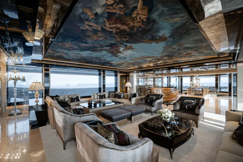 Luxurious yacht interior lounge with painted celestial ceiling, plush seating, lamps, and ocean views through large windows.