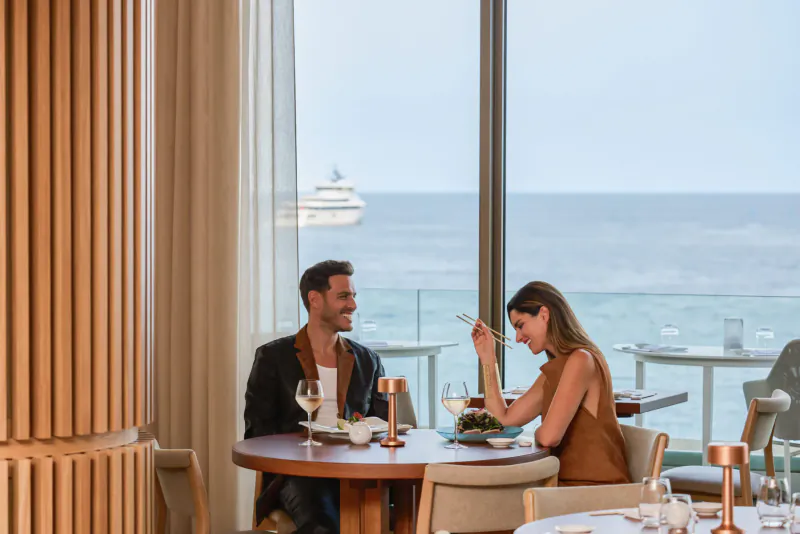 Smiling couple dining at round table in elegant restaurant with ocean view and yacht beyond large windows