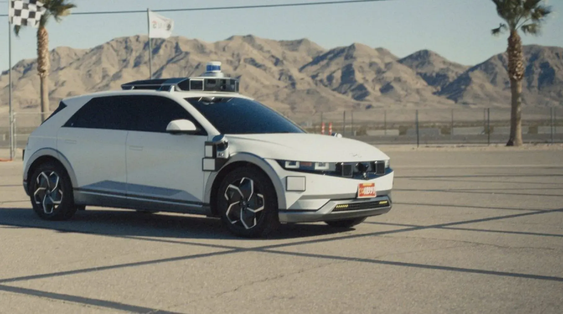 White Hyundai IONIQ 5 robotaxi with roof lidar passes DMV driving test on desert lot with palms and mountains.