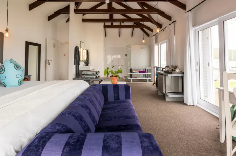 Spacious loft room at The Lofts Boutique Hotel with purple striped sofa, white bed, kitchenette, and wooden beams overlooking lagoon.