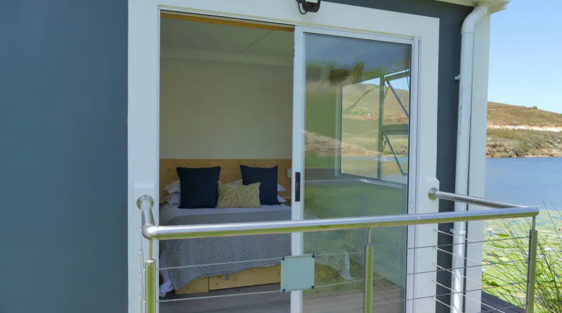 Open sliding door to cozy bedroom with gray bed and pillows, overlooking lake and hills from balcony in Elgin Valley.