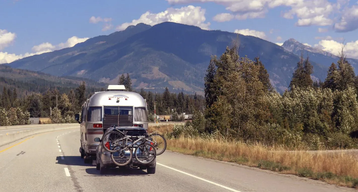 Silver campervan with bike rack towing bikes on highway amid mountains and pine trees under blue sky.