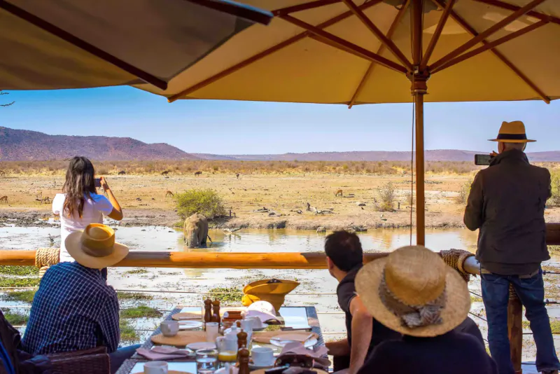 Group at outdoor lodge table under umbrellas, photographing savanna landscape with river at Tau Game Lodge