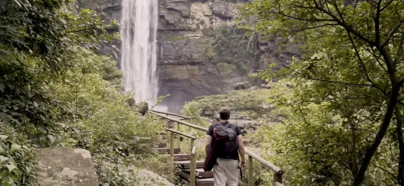Hiker with backpack walks up wooden steps toward lush tropical waterfall surrounded by greenery