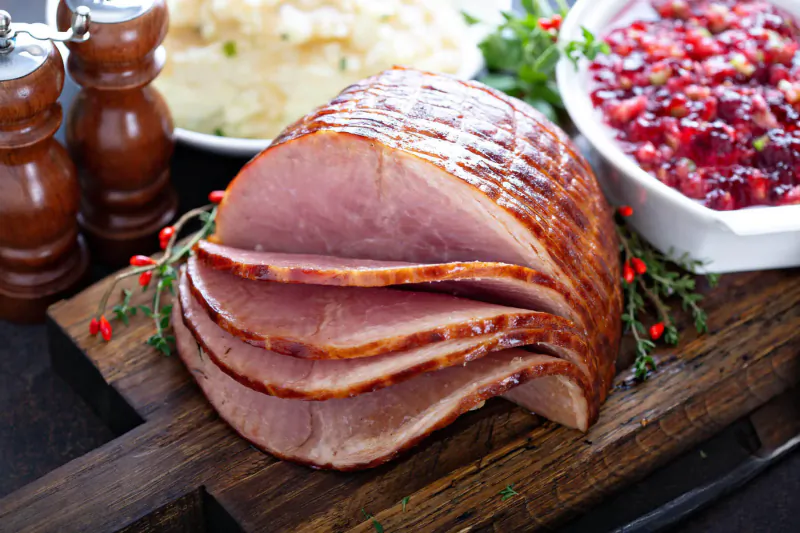 Sliced glazed ham on wooden board with mashed potatoes, cranberry sauce, garnished with herbs and pepper mills