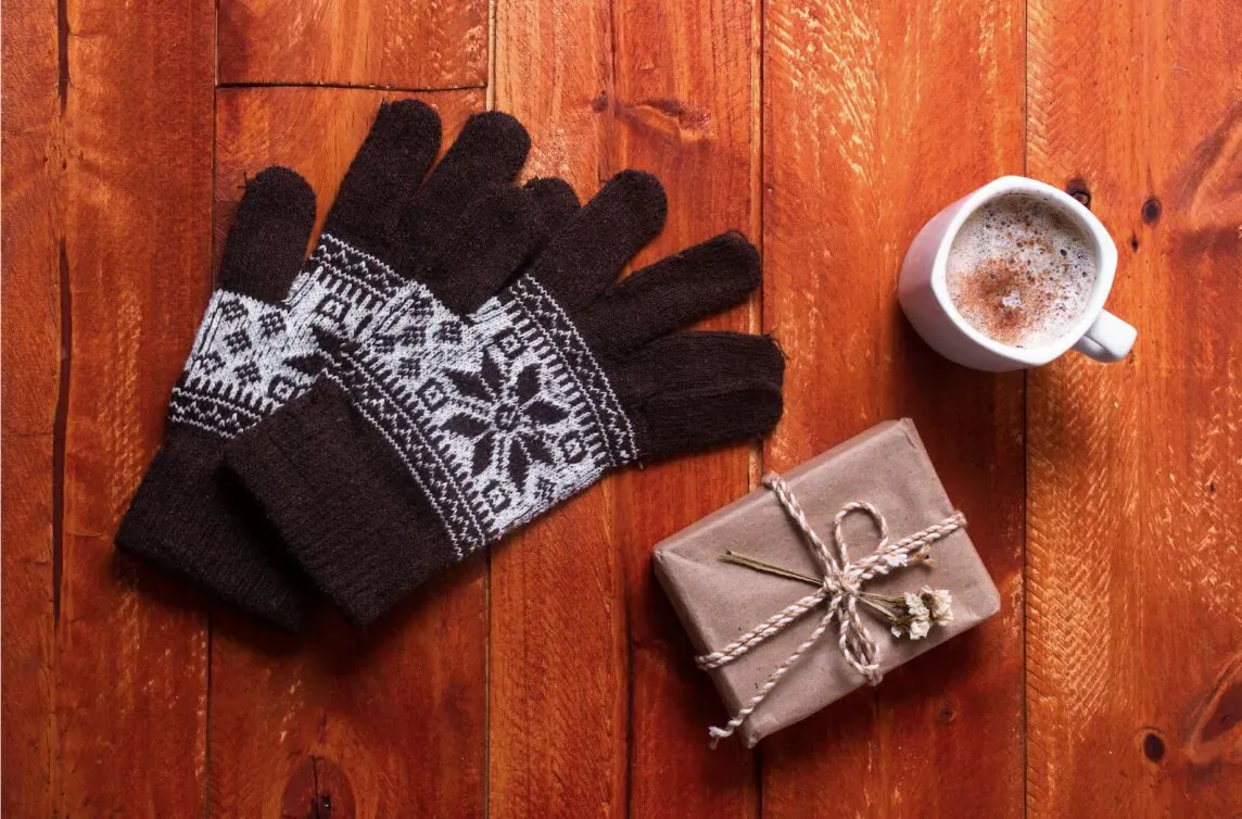 Brown knitted Nordic gloves with snowflake patterns beside tied gift box and hot cocoa mug on wooden table.