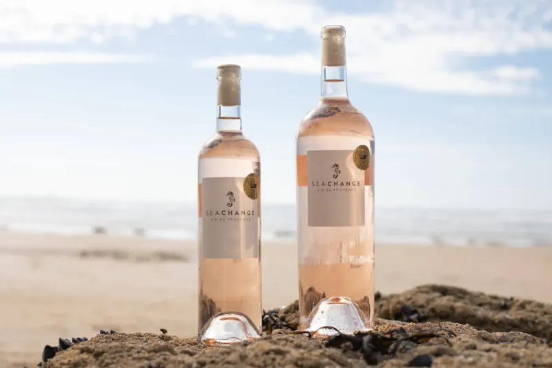 Two bottles of Sea Change rosé wine on beach rocks by ocean under blue sky