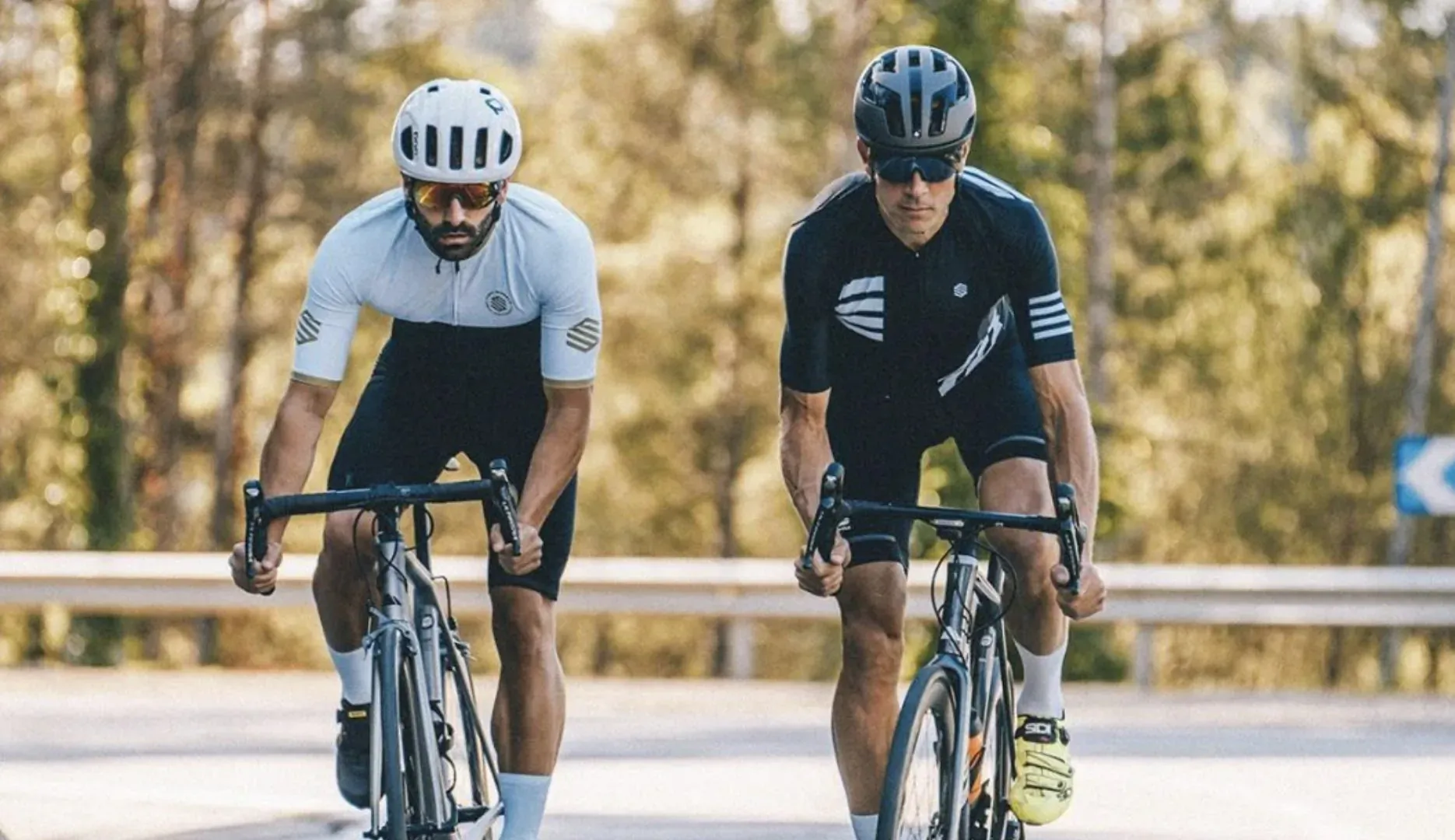 Two cyclists in helmets and jerseys ride road bikes side-by-side on a forested mountain road.