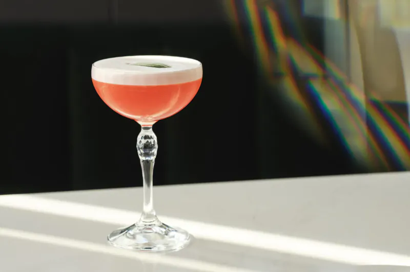 Pink cocktail with white foam in elegant coupe glass on white table, rainbow light prism and soft shadows, rooftop bar vibe.