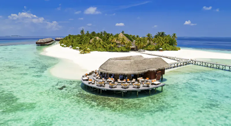 Aerial view of thatched-roof overwater restaurant on Mirihi Island Resort, surrounded by turquoise lagoon, white sand, and palms.