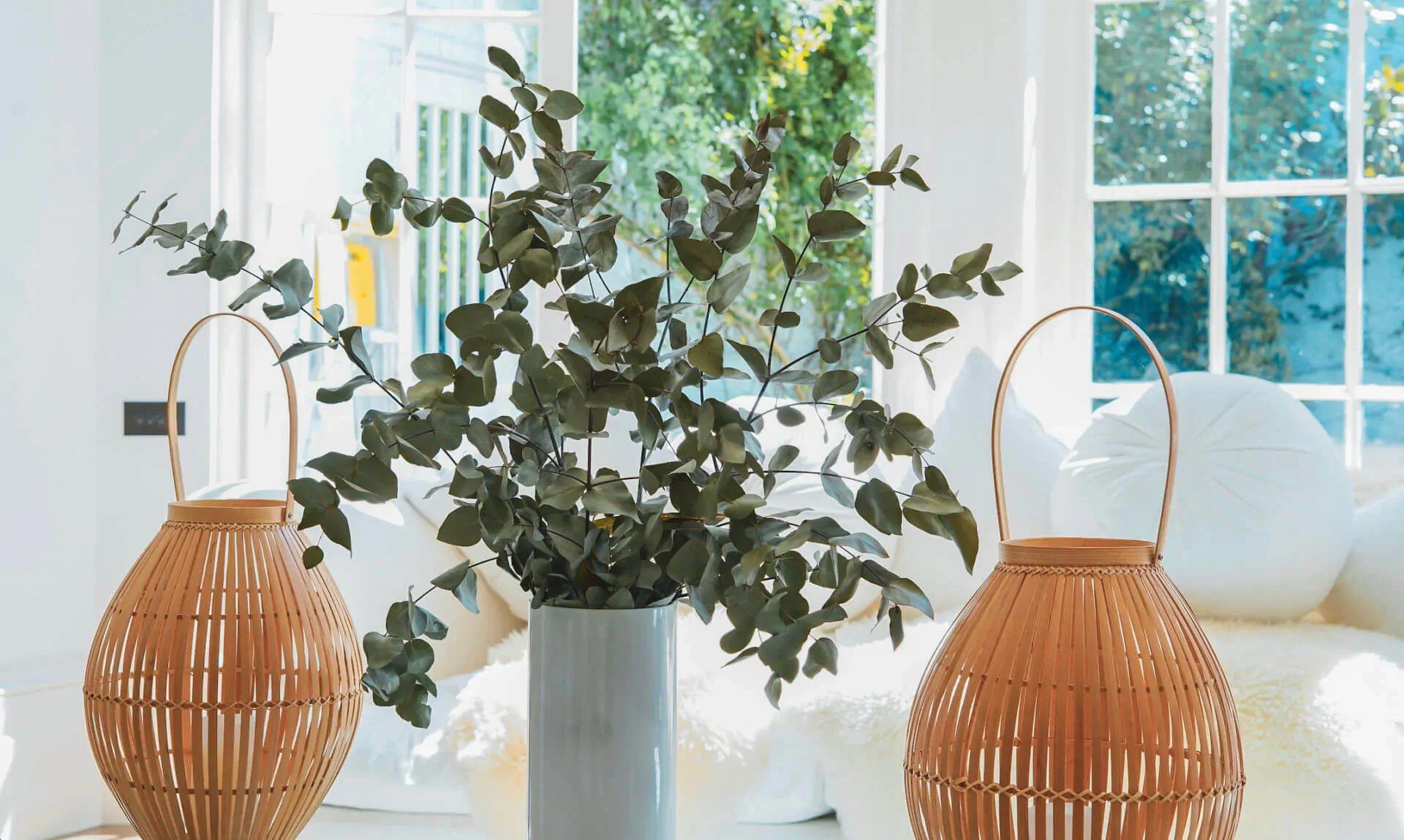 Two rattan lanterns flanking eucalyptus in vase on white sofa in sunlit luxurious modern interior