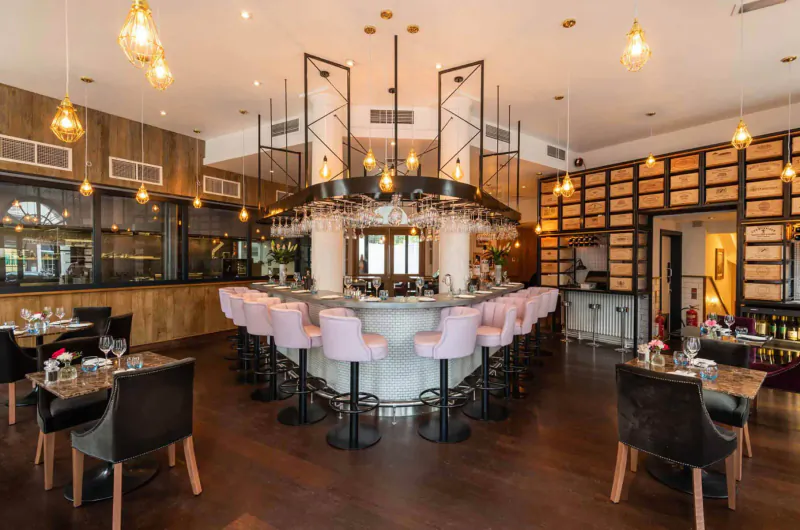 Elegant 28°-50° Chelsea bar with curved white counter, pink stools, wood walls, pendant lights, and tables.