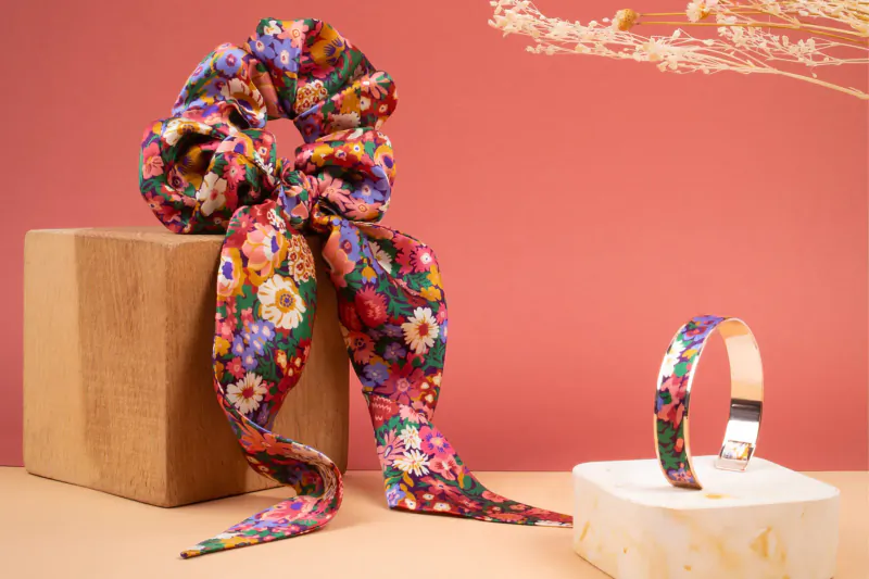 Colorful floral scrunchie on wooden box with matching bangle on white stand, pink backdrop with dried grass.
