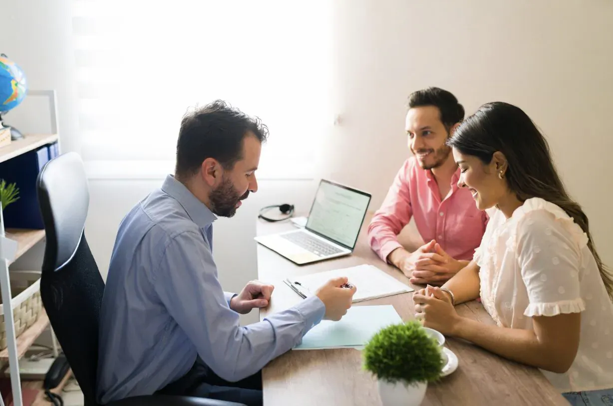 Male advisor in blue shirt discusses tax documents with smiling couple at office desk with globe and plant