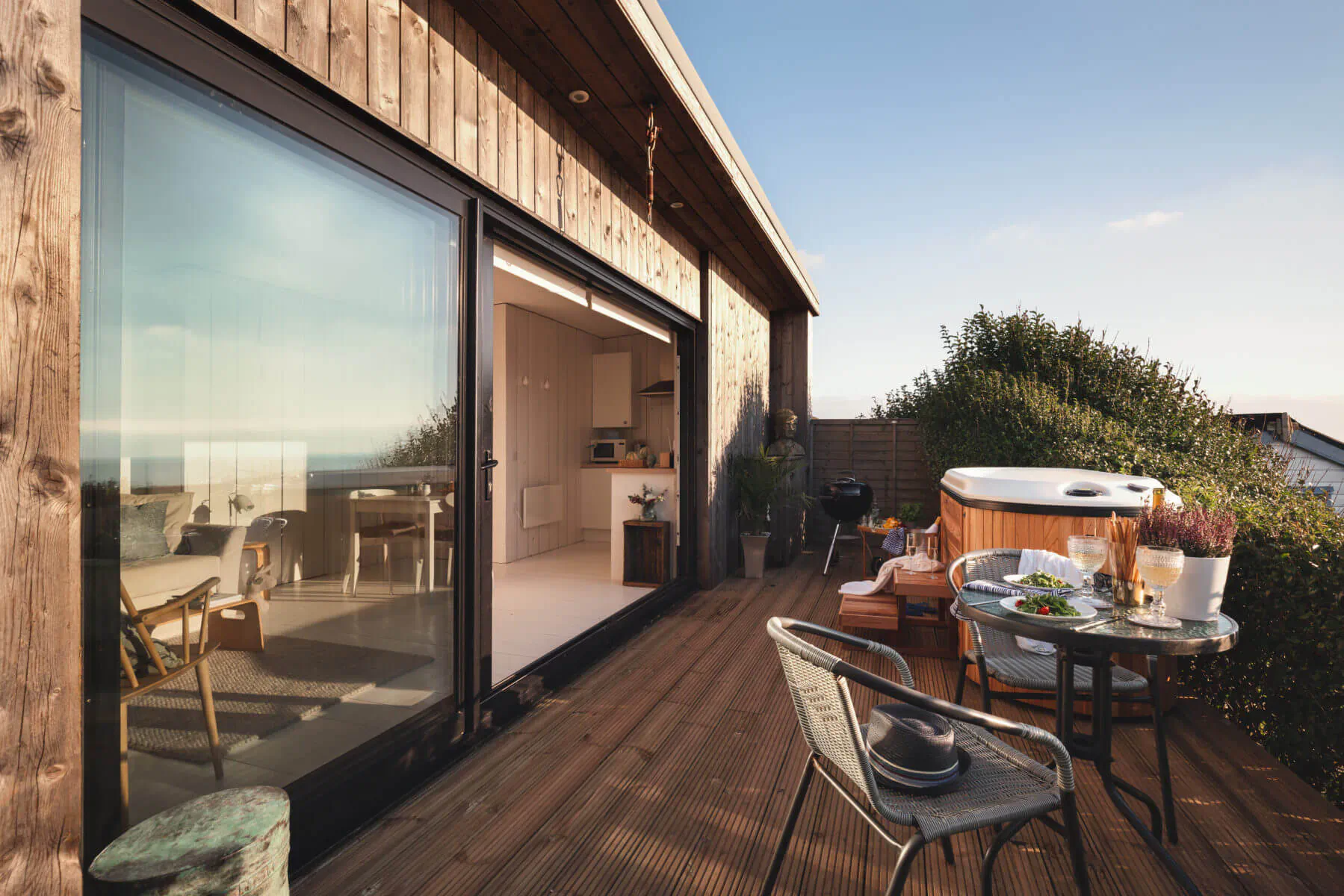 Modern wooden cabin deck with open glass doors to kitchen, hot tub, chairs, table, plants, ocean view at sunset