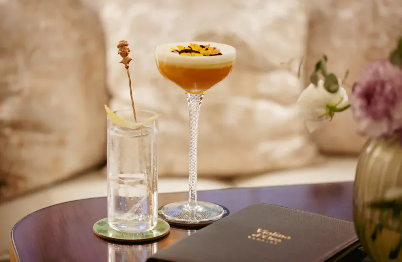 Elegant cocktail with orange foam and flowers on a low table beside a water glass at Violino d'Oro Hotel, Venice.