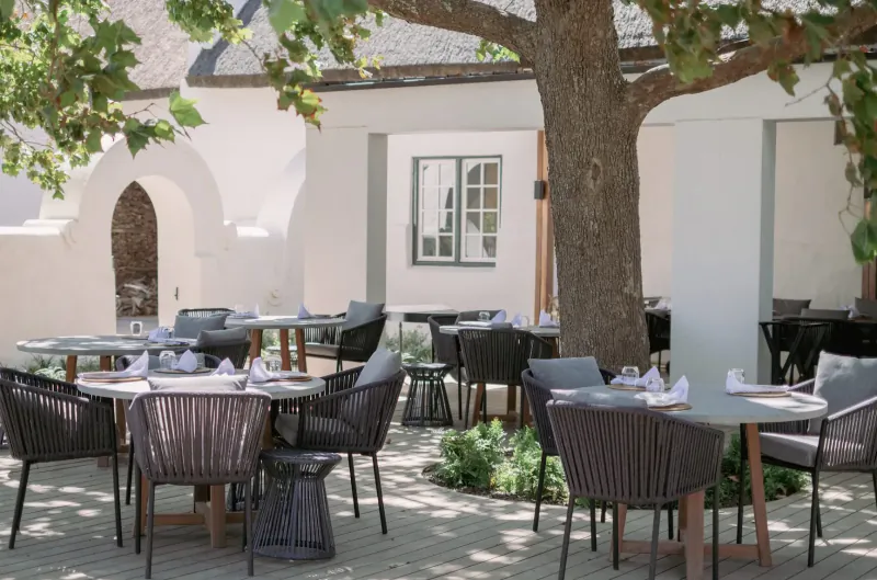 Outdoor dining area at Clara’s Barn, Vergenoegd Löw: wicker tables with white napkins under large tree by white adobe walls.