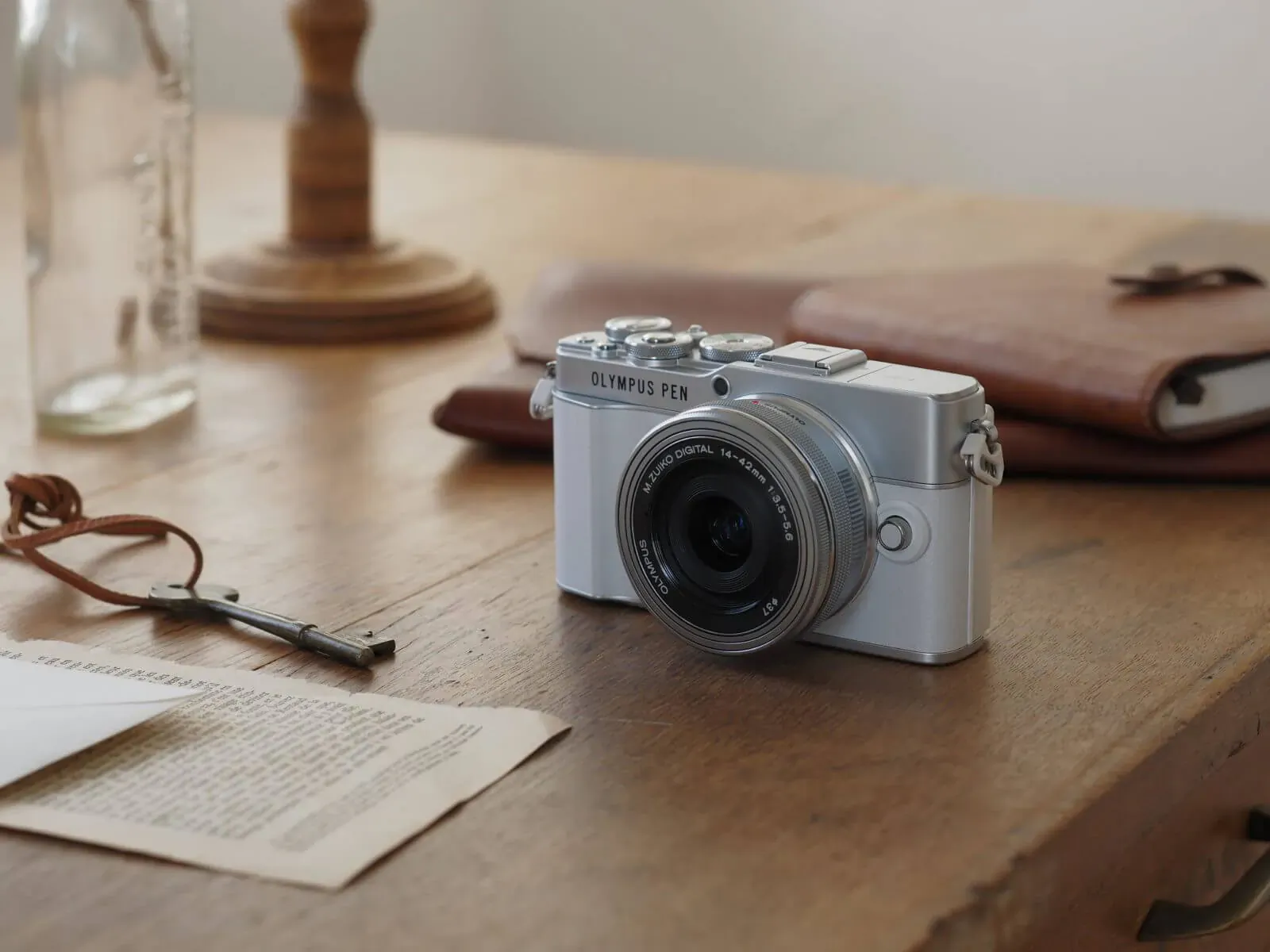 Silver Olympus PEN E-P7 camera on wooden desk with leather notebook, glasses, bottle, and papers.