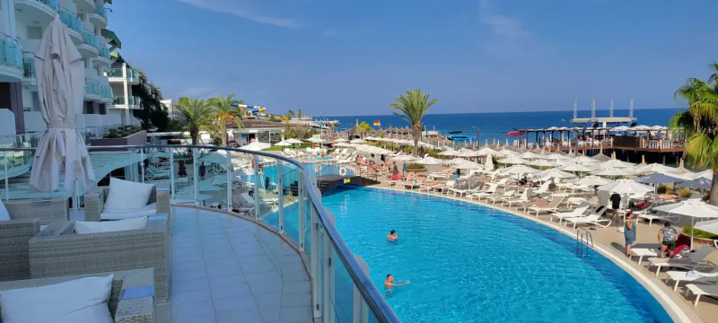 Aerial view from luxury hotel balcony over curved infinity pool with swimmers, white loungers, beach, sea, and palm trees at Corendon Playa Kemer, Turkey.
