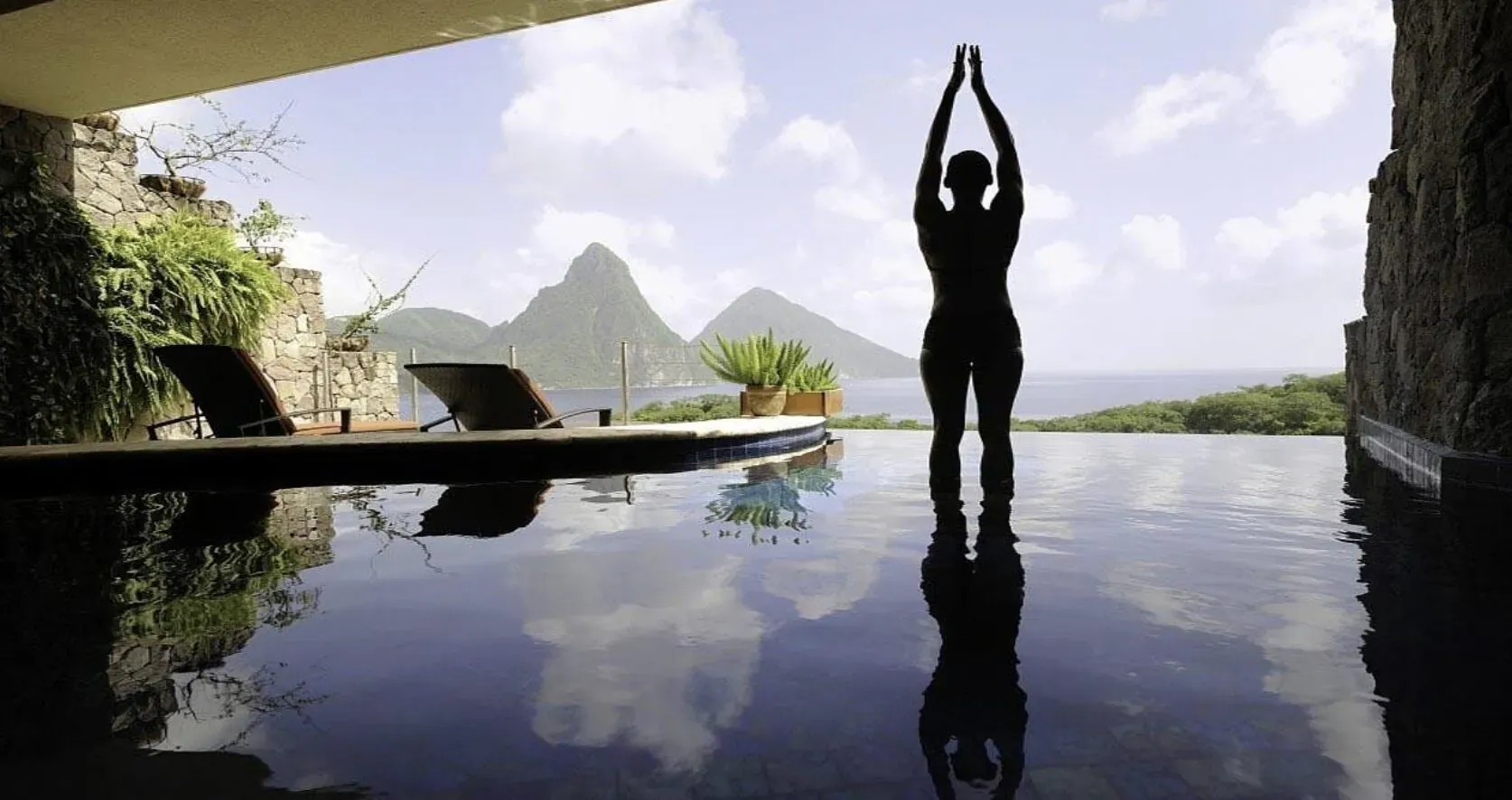 Silhouette of woman in yoga pose at infinity pool edge, Pitons mountains and ocean view, wellness retreat.