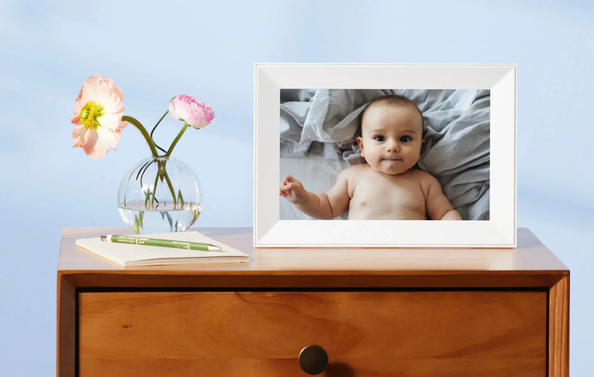 Aura smart frame on wooden dresser showing smiling baby in crib, with pink flowers in vase beside it.
