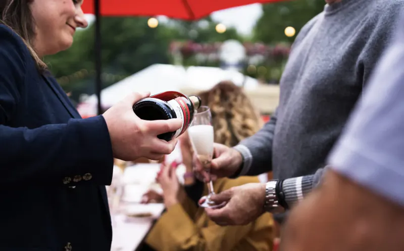Woman in suit pours champagne from bottle into flute held by man at outdoor Taste of London event under red umbrella.