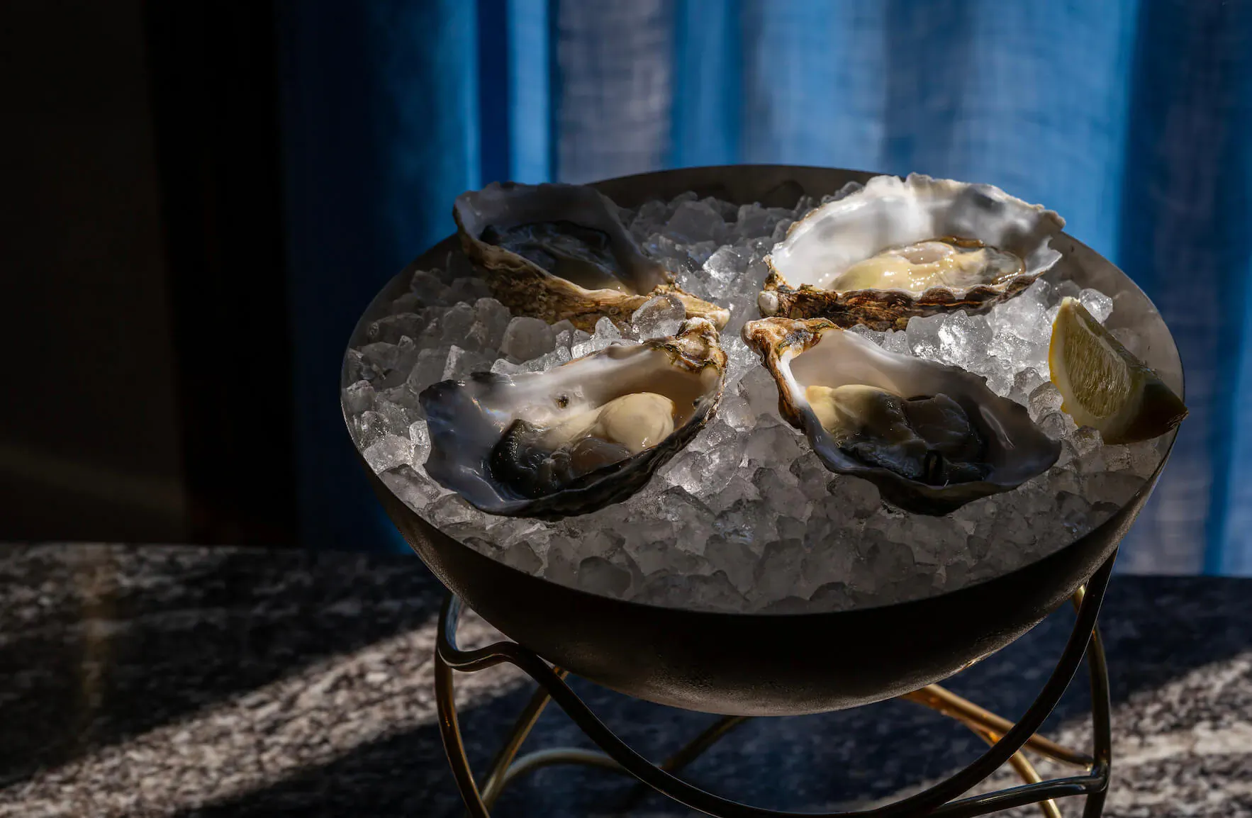 Fresh oysters on ice in a gold metal bowl with lemon wedges, on marble surface against blue curtain.