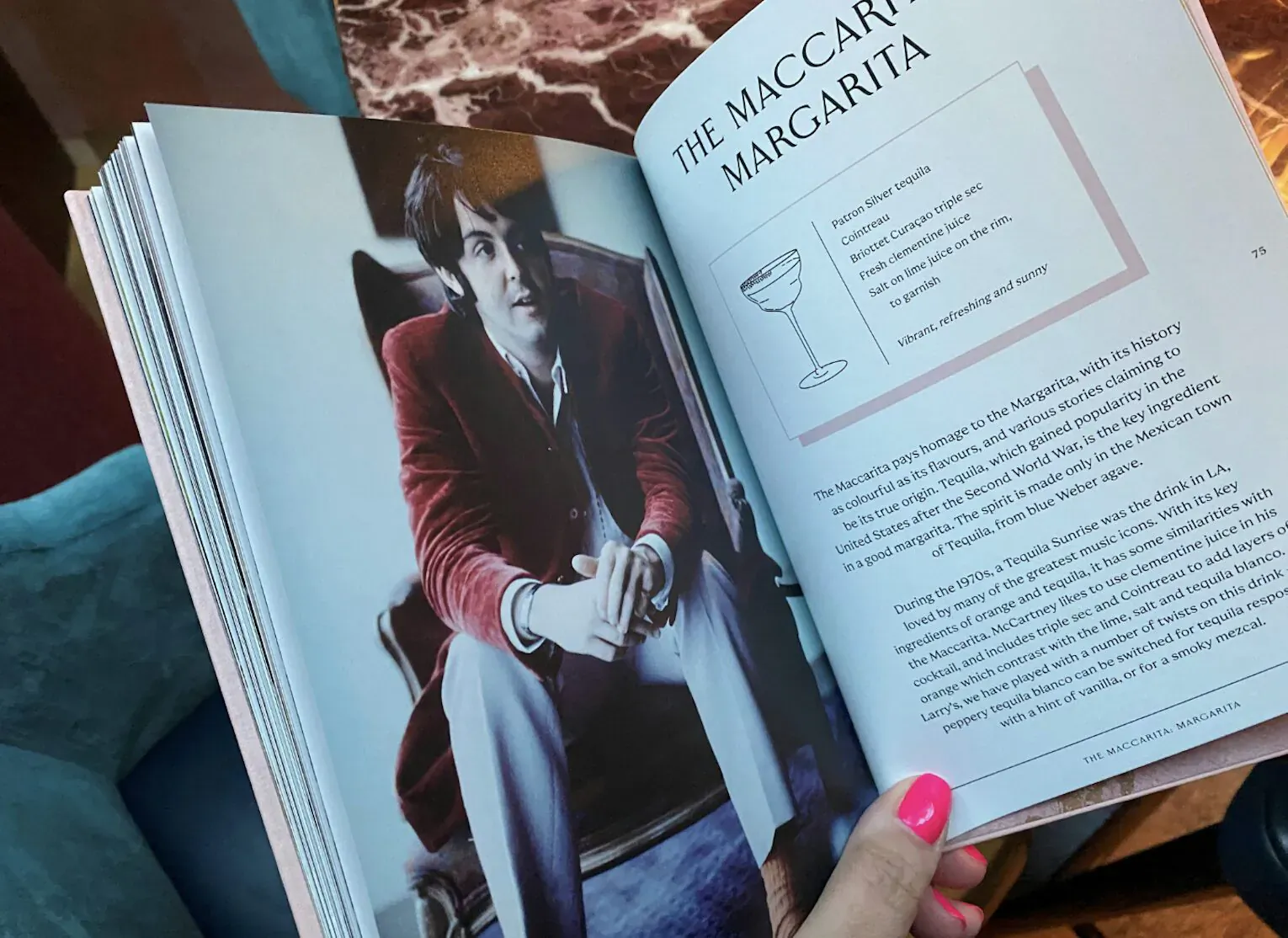 Open cookbook page 'The Margarita' recipe with cocktail glass illustration, held by manicured hand over man in red velvet jacket