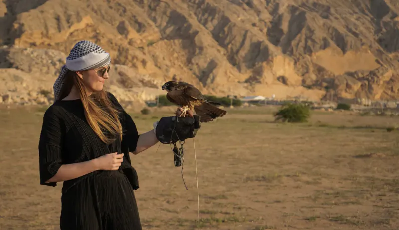 Woman in black dress and headscarf holds falcon on gloved hand in desert with sandy mountains behind