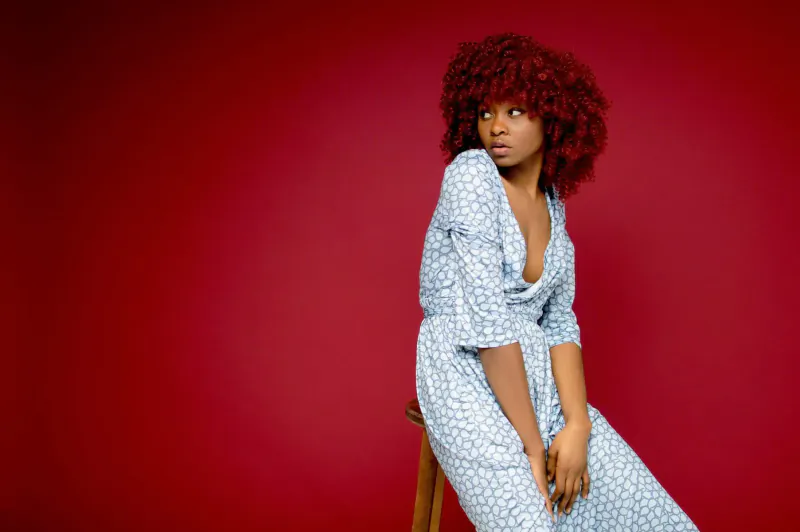 Woman with curly red hair sits sideways on wooden stool in white floral dress against red background