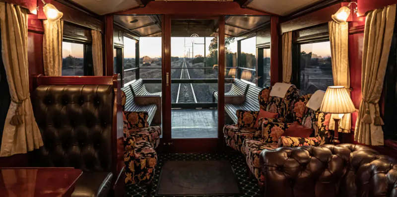 Luxurious train lounge with floral sofas, lamps, large windows showing railway tracks at sunset.