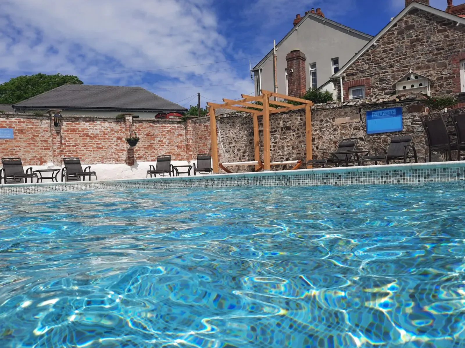 Close-up of shimmering blue pool at Broomhill Manor, with lounge chairs, pergola, brick walls, and stone buildings under partly cloudy sky.
