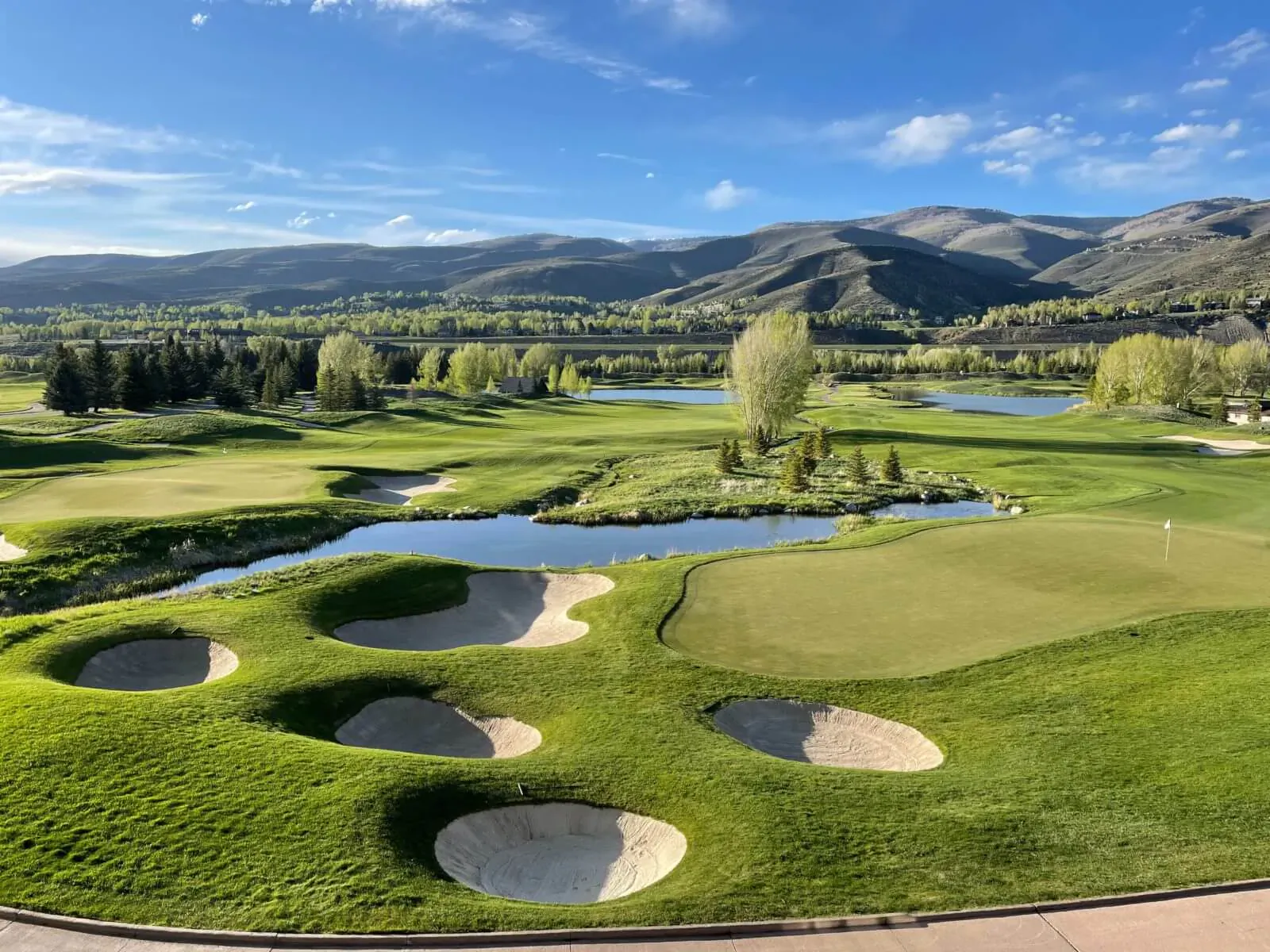 Aerial view of lush green golf course with bunkers, ponds, trees, and distant mountains under blue sky.