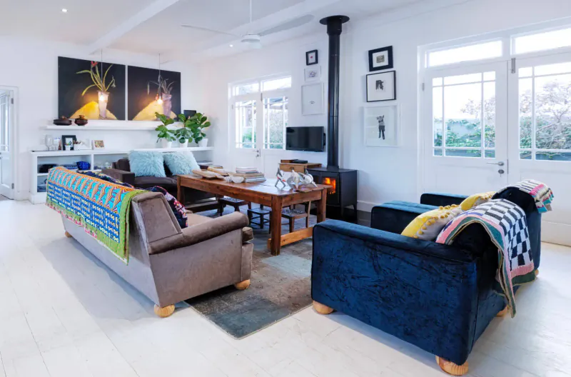 Spacious living room in Vishuis Cottage with brown and blue sofas, wooden table, black wood stove, art on white walls, large windows.