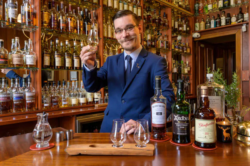Smiling man in blue suit holds whisky glass at wooden bar with Glendronach, Glenfiddich bottles and tasting glasses.