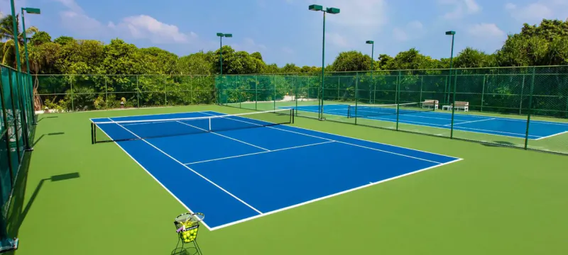 Two blue tennis courts with green padding, surrounded by mesh fences and lush tropical trees at Hideaway Beach Resort, Maldives.
