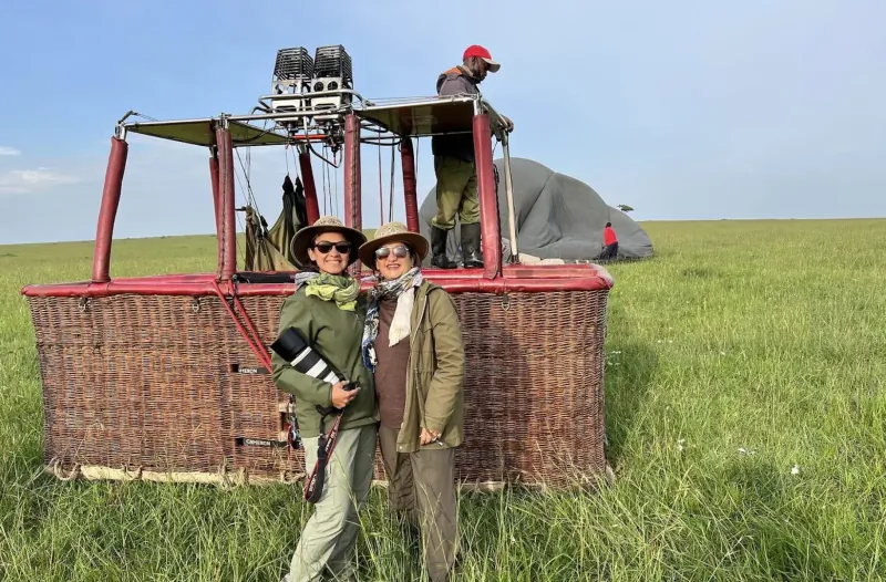 Two smiling adventurers in safari gear stand by a red hot air balloon on African savanna grassland.