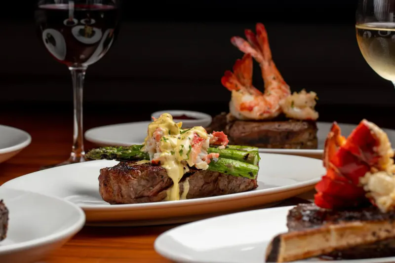 STK Steakhouse plate of seared ribeye steak topped with lobster, asparagus, and sauce beside wine glasses