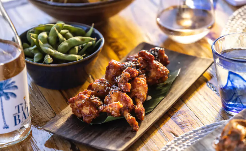 Glazed chicken wings on banana leaf with edamame, rosé wine 'The Bay', on wooden table at Rokuni Antigua.