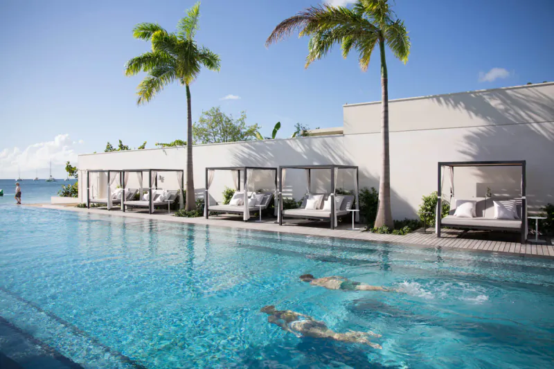 Silversands Grenada infinity pool with swimmers, white cabanas, palm trees, and beachside building under blue sky.
