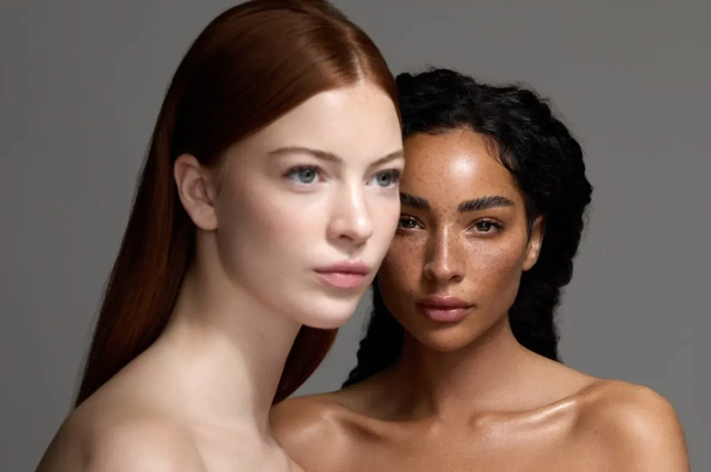Close-up portrait of two women with flawless skin: redhead and Black woman with curly hair, gazing intently on gray background