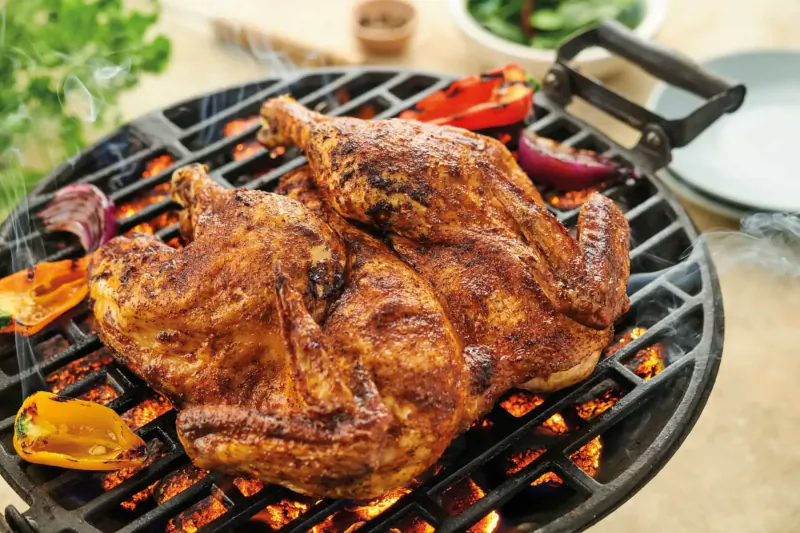 Two seasoned chicken thighs grilling on a black barbecue with peppers, garnished with greens