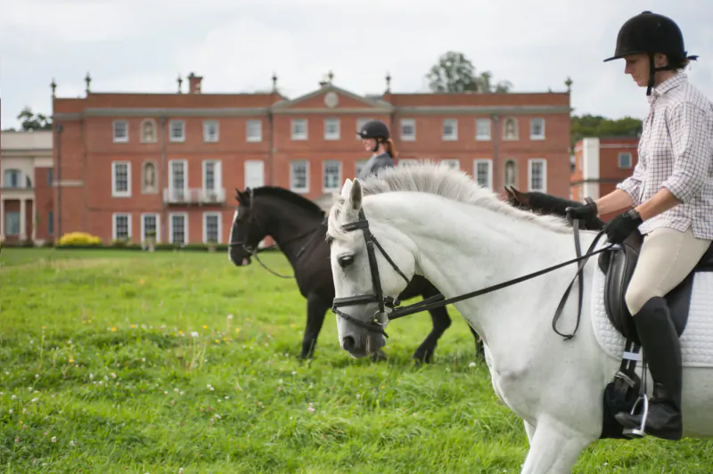 Woman in riding gear on white horse with black horse beside, in front of grand red brick manor at Four Seasons Hampshire.