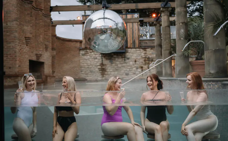 Five women in swimsuits smiling and toasting drinks in a stylish hot tub at The Rabbit Hotel & Retreat, with disco ball and stone ruins backdrop.